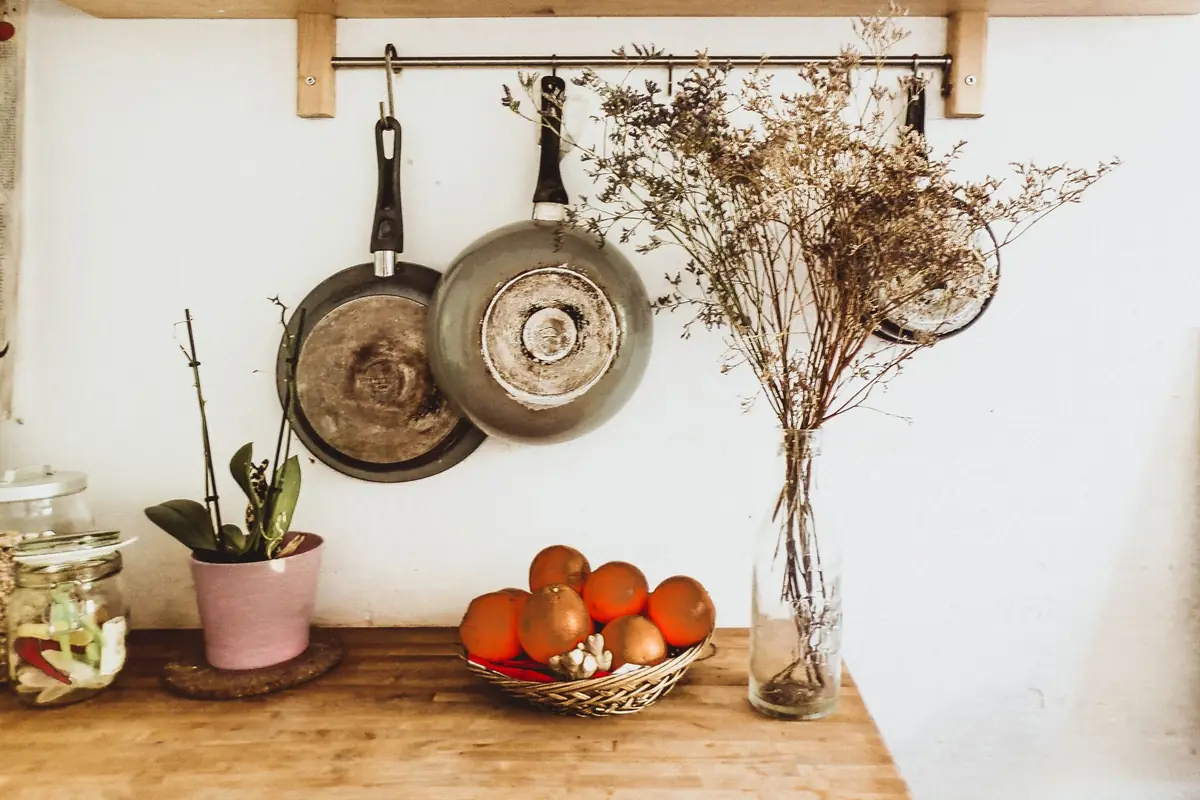 Two frying pans hanging from a wall rail above a wooden kitchen counter with a bowl of oranges